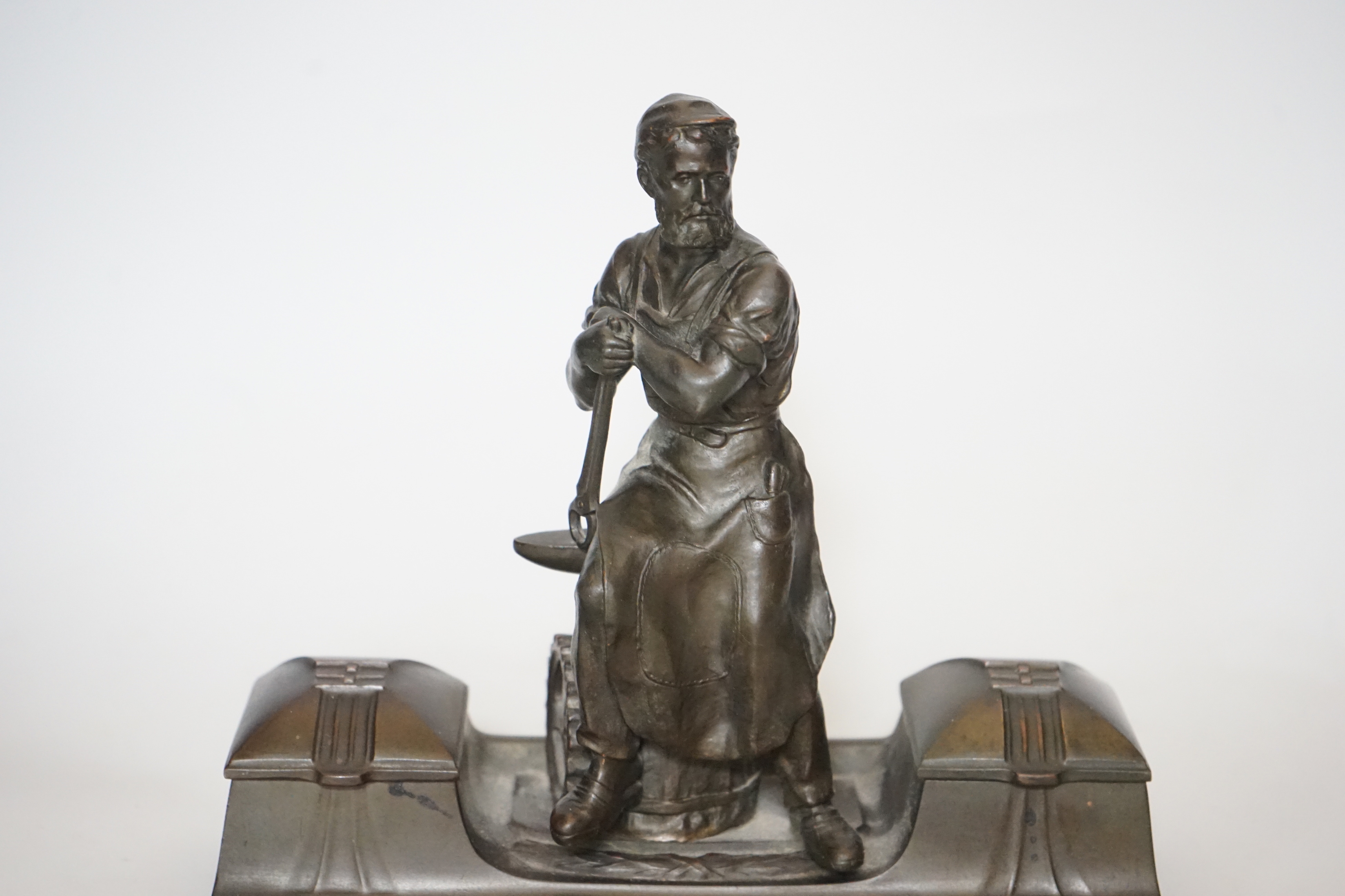 A WMF style spelter ‘Industry’ inkstand, figure with tools and an anvil, hinged ink wells either side, one glass ink bottle present, ‘1265’ cast into base, 20cm high